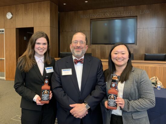 From left to right, Julia, Bennett, and Victoria stand in suits smiling. They are standing in a lecture room at American University with woodpaneled walls. Julia and Victoria are each holding a Bad Spaniels dog toy